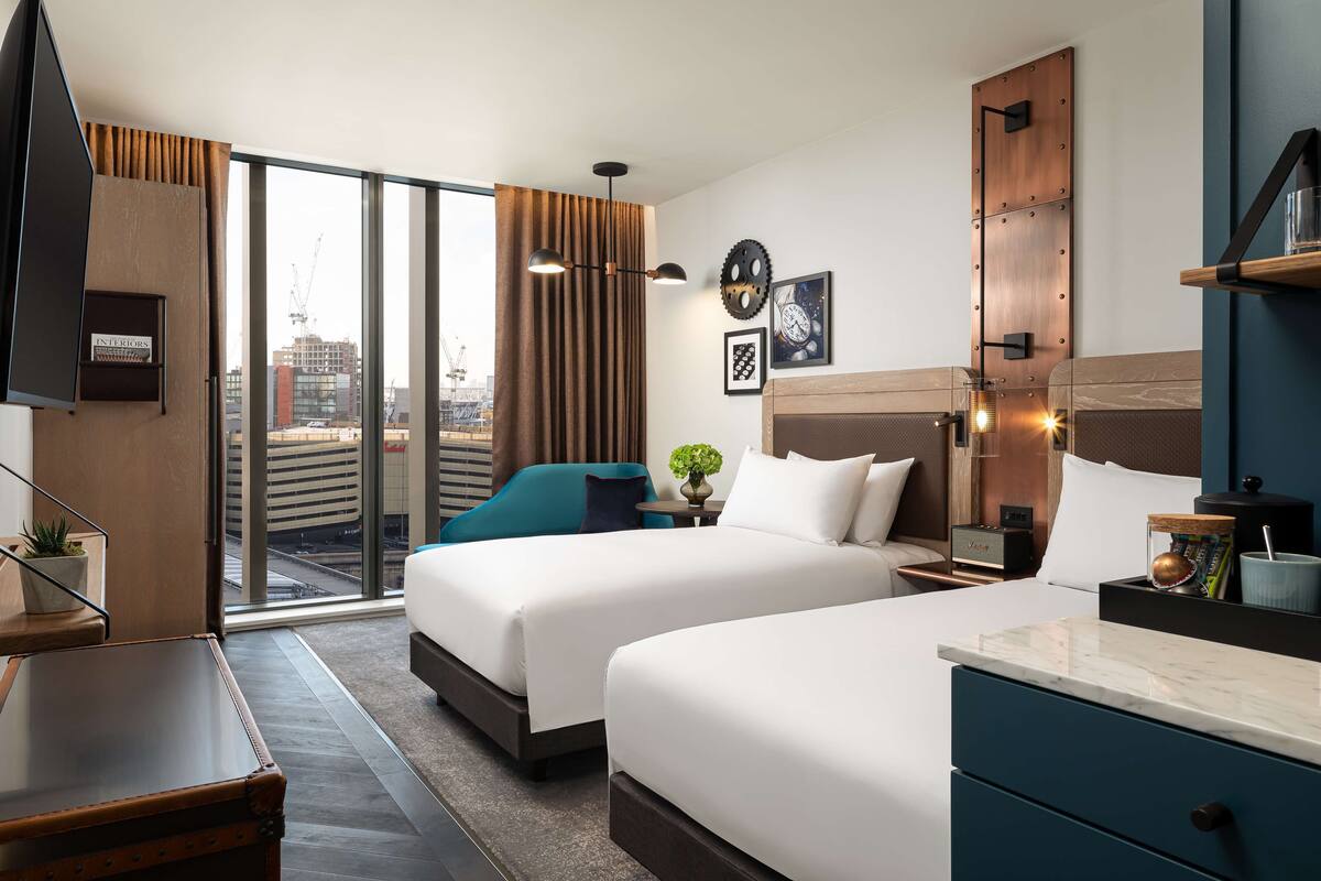 A hotel room with two beds, a desk, a TV, a large window with a view of the city, and a modern decor style.