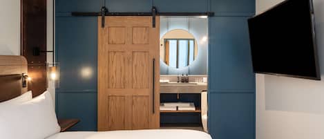 A hotel room with a bed, a wooden door, a bathroom with a mirror, and a mounted TV.