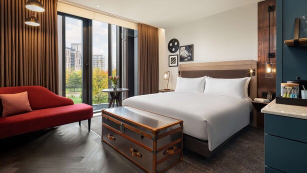 A hotel room with a large bed, a red sofa, a wooden chest of drawers, and a view of the city through large windows.