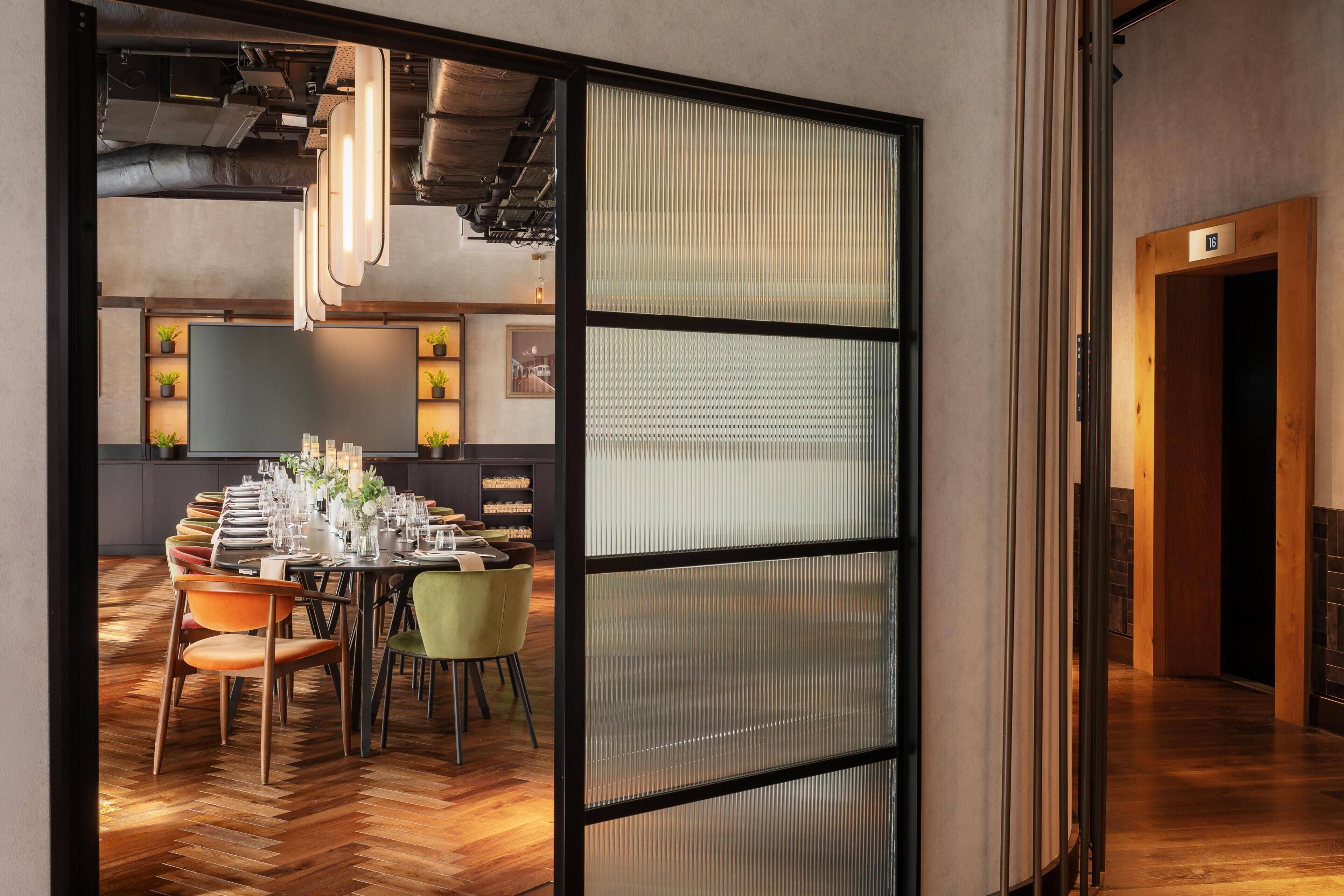 Un comedor moderno con una larga mesa dispuesta para una comida, rodeada de sillas, y vistas a una zona de cocina con estantes y plantas.
