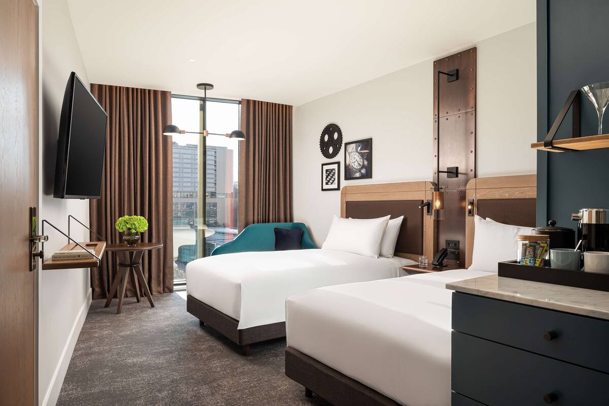 A hotel room with two beds, a TV, a desk with a plant, a window with curtains, and a city view.