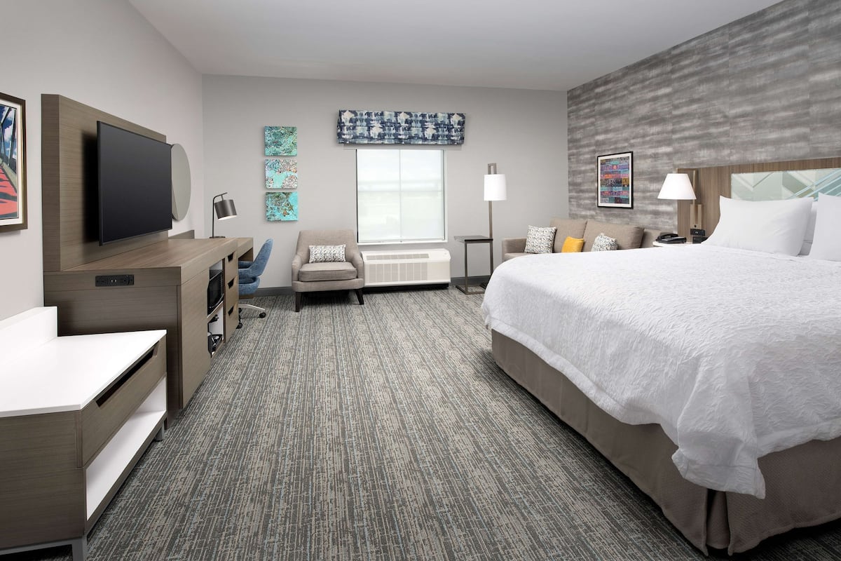 A hotel room with a large bed, a desk with a flat-screen TV, a chair, and a window with blinds.