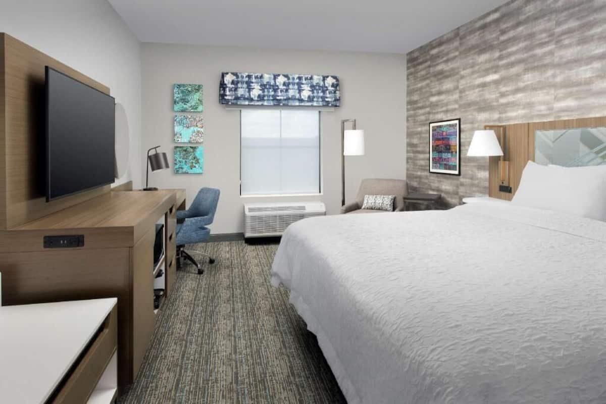 A hotel room with a bed, desk, chair, TV, artwork, and a window with blinds.