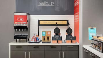 Una estación de desayuno con dispensador de jugo, máquina para hacer gofres y área para la masa de gofres.