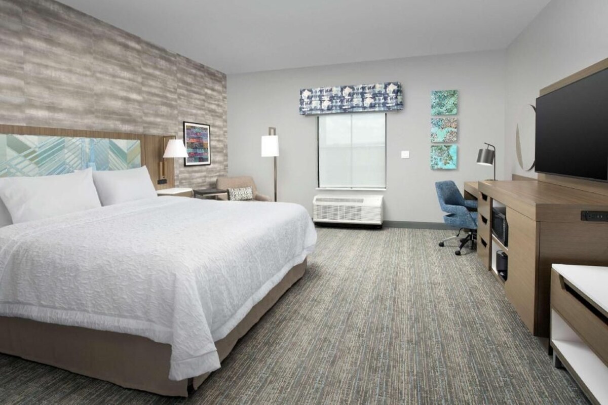 A hotel room with a bed, a desk, a chair, a flat-screen TV, a window, and artwork on the walls.