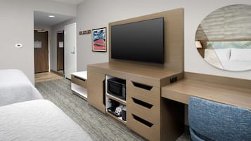 A hotel room with a large flat-screen TV mounted on a wooden cabinet, a bed with white bedding, a blue upholstered chair, and a framed artwork on the wall.