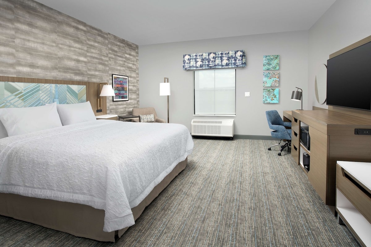 A hotel room with a large bed, a desk with a computer, a chair, an air conditioning unit, and artwork on the walls.