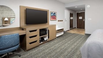 A hotel room with a flat-screen TV, a wooden desk, a blue office chair, a microwave, a coffee maker, a large mirror, and a bed with white bedding.