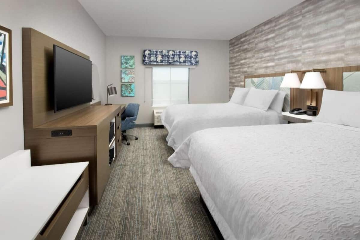 A hotel room with two beds, a desk, a TV, and a window.