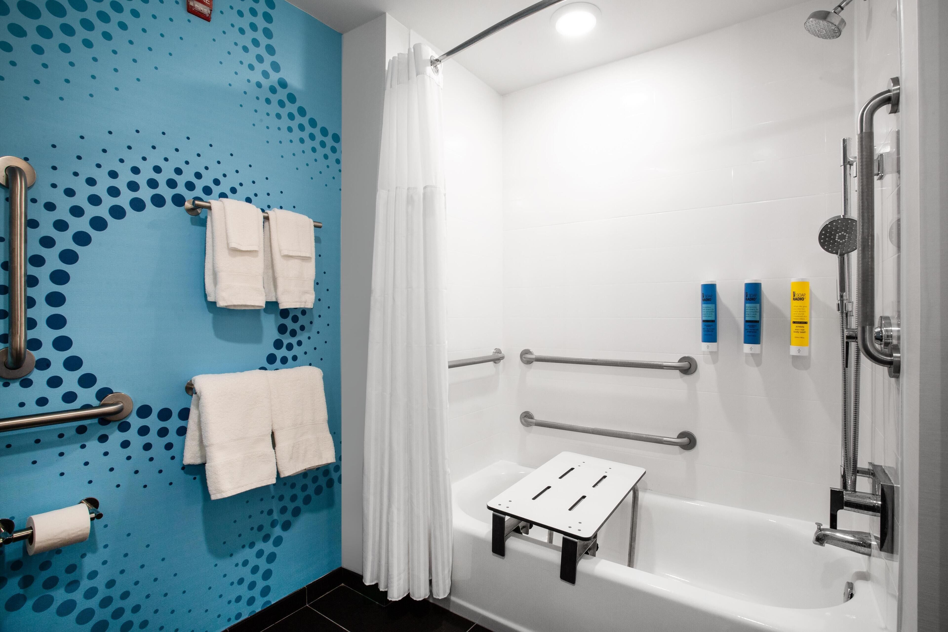 A modern bathroom with a blue wall, white tiles, a shower with a handheld showerhead, a grab bar, and a small step stool.