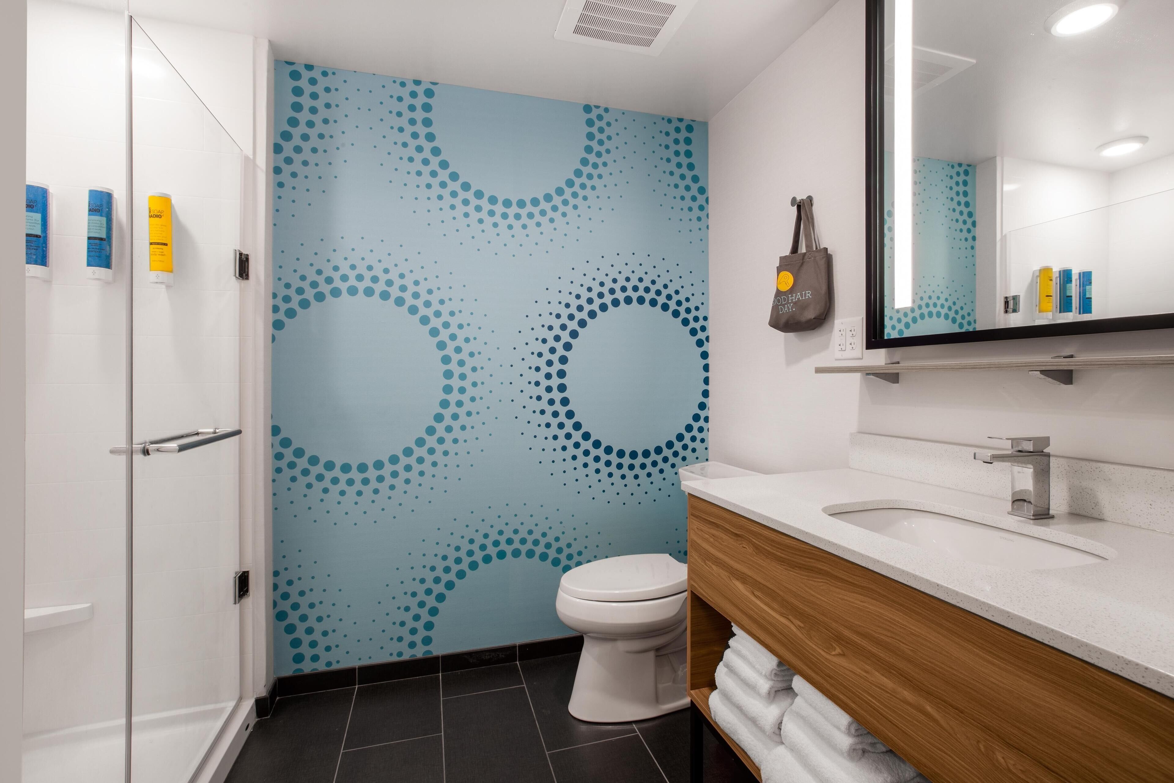 A modern bathroom with a blue patterned wall, a white toilet, a wooden vanity, and a mirror above the sink.