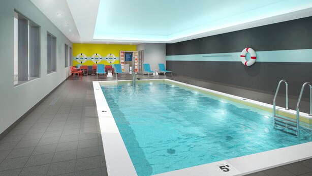 A modern indoor swimming pool with clear water, a lifebuoy on the wall, and a seating area with chairs and tables.