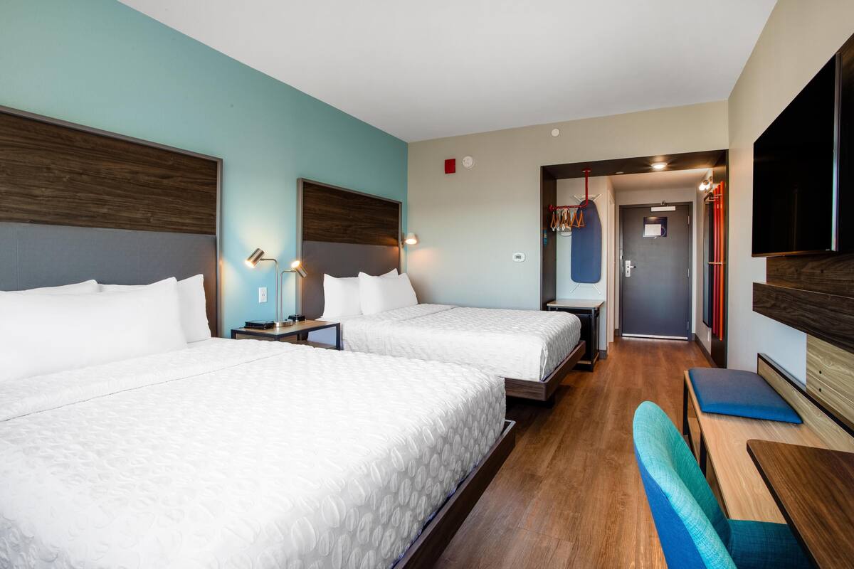 A hotel room with two beds, wooden headboards, a bench, a TV, and a door leading to a hallway.