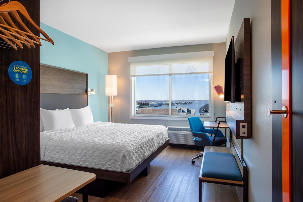 A hotel room with a bed, desk, chair, and a large window offering a city view.