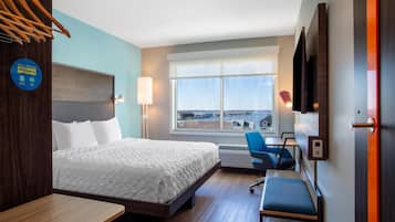 A hotel room with a bed, desk, chair, and a large window offering a city view.