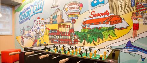 A room with a foosball table, a mural depicting a coastal city scene, and a red ottoman.