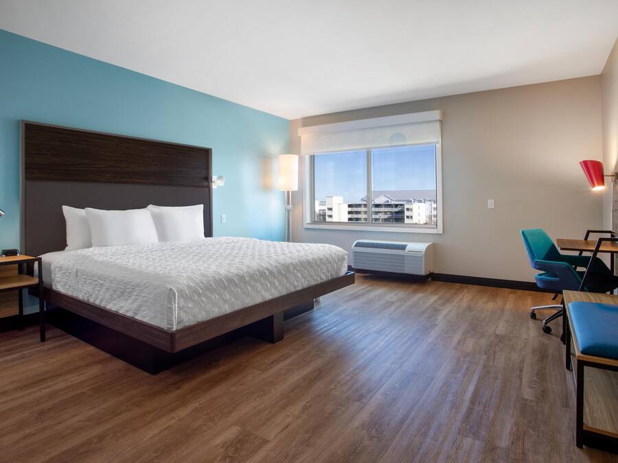 A hotel room with a large bed, a desk, a chair, a window with a view, and an air conditioning unit.
