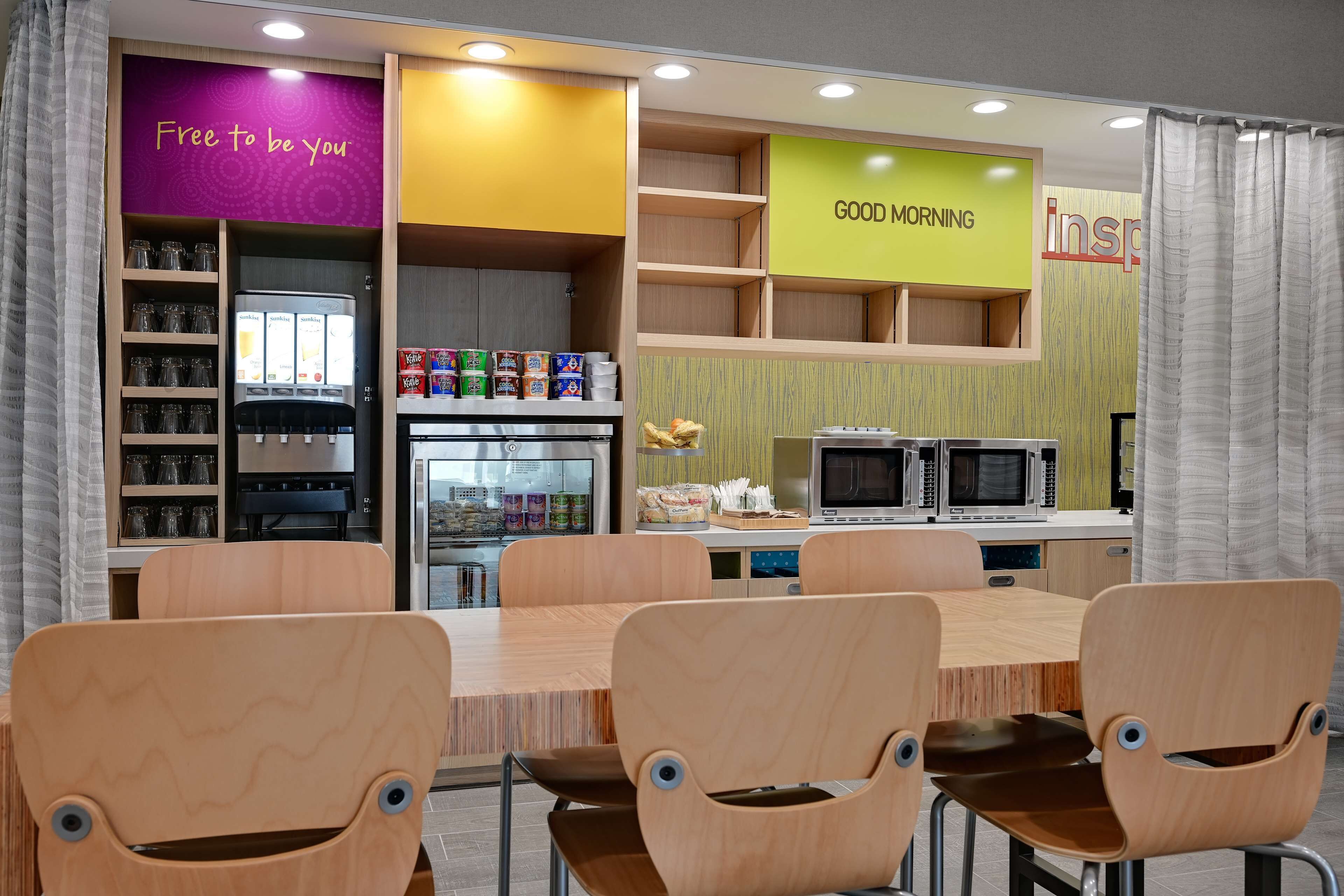 A modern kitchen with wooden cabinets, a refrigerator, microwave, and a coffee machine. There are wooden chairs around a table and a shelf with snacks.