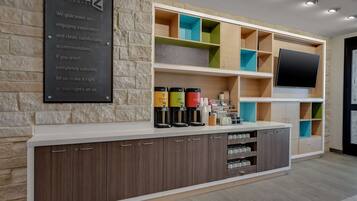 A reception area with a stone wall, a signboard, a counter with coffee dispensers, and a TV mounted on the wall.