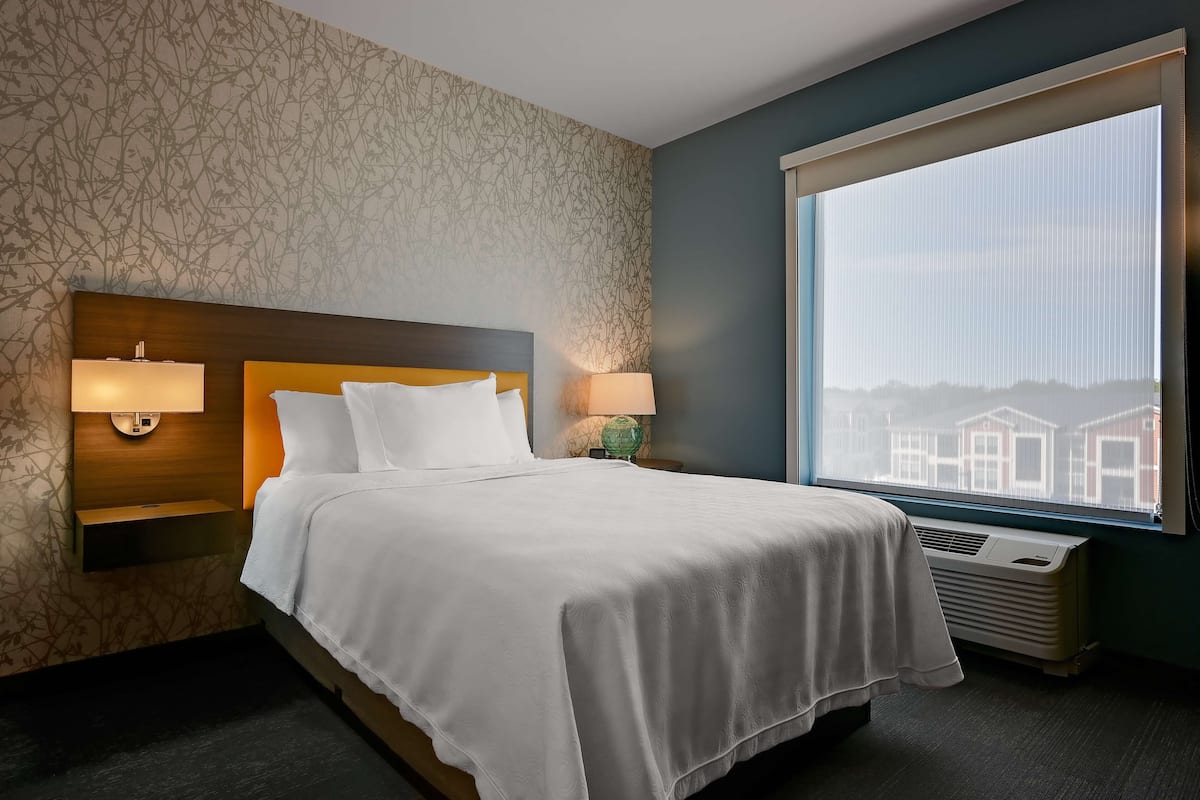 Habitación de hotel con un ventanal grande, cama con ropa blanca, mesita de noche con lámpara y un calefactor fijado a la pared.