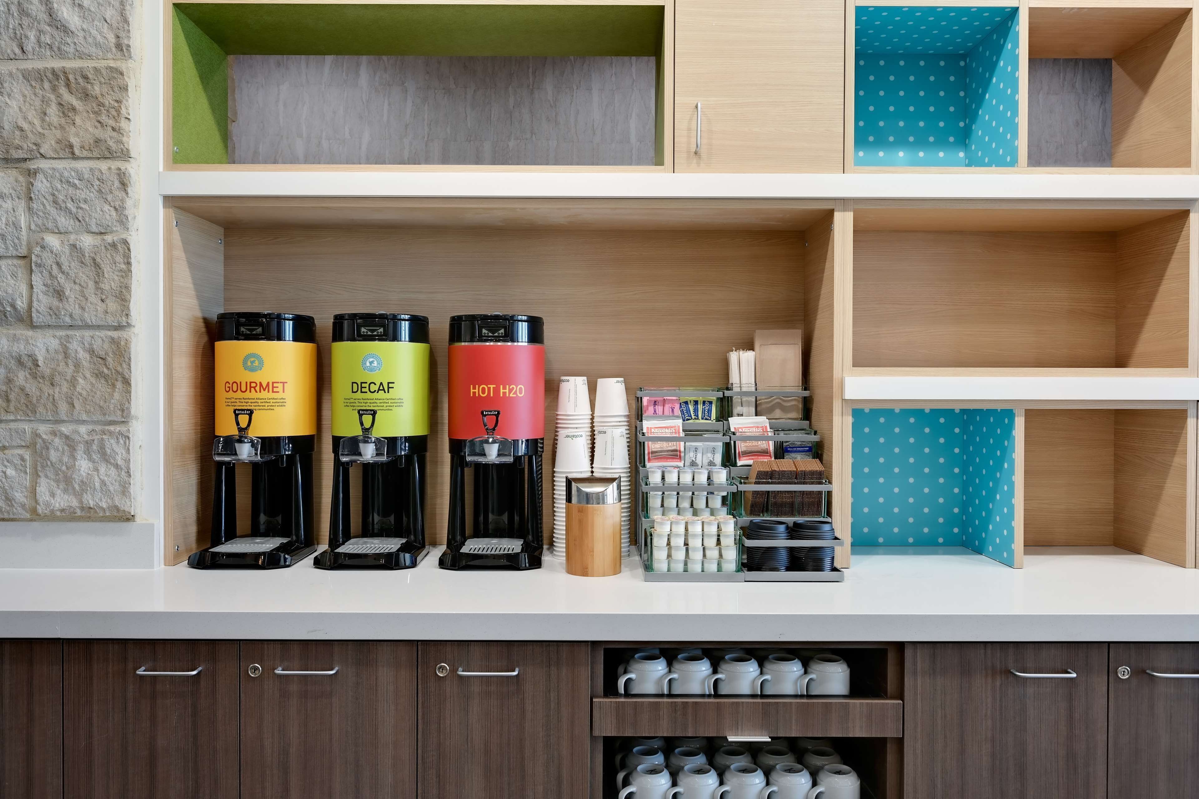 A coffee station with three coffee makers labeled 'GOURMET,' 'DECAF,' and 'HOT H2O.' There are also stacks of white cups and a variety of coffee pods.