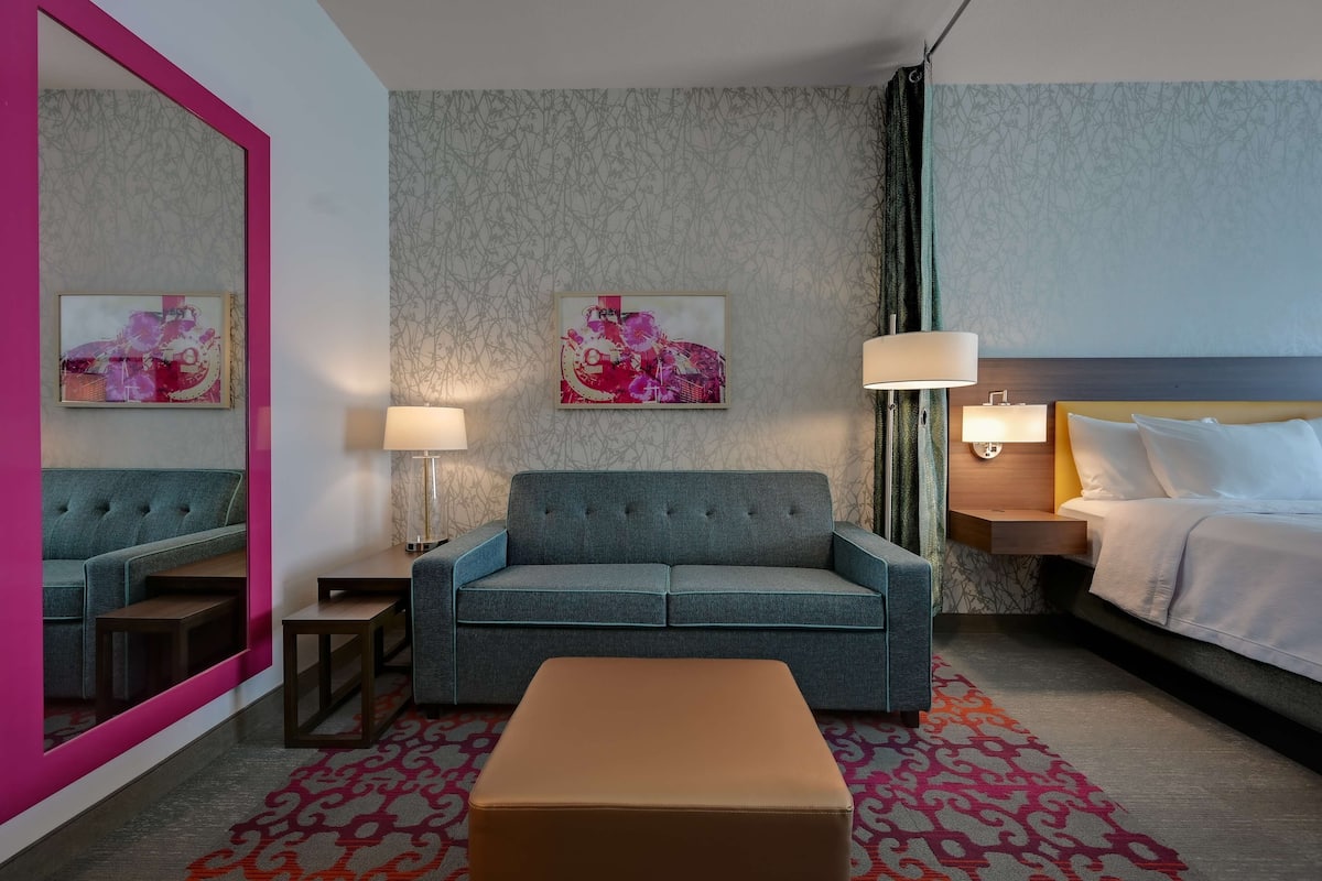 Habitación de hotel con sofá azul, cama con ropa de cama blanca, un espejo grande, dos mesitas de noche con lámparas y una alfombra estampada.