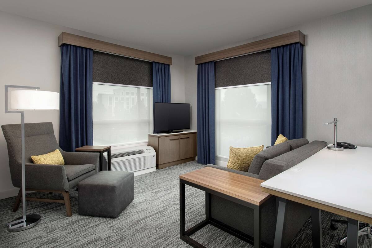 Une chambre d’hôtel moderne avec un canapé gris, une table basse en bois, un fauteuil gris, un téléviseur posé sur un meuble en bois et une petite table avec une lampe.