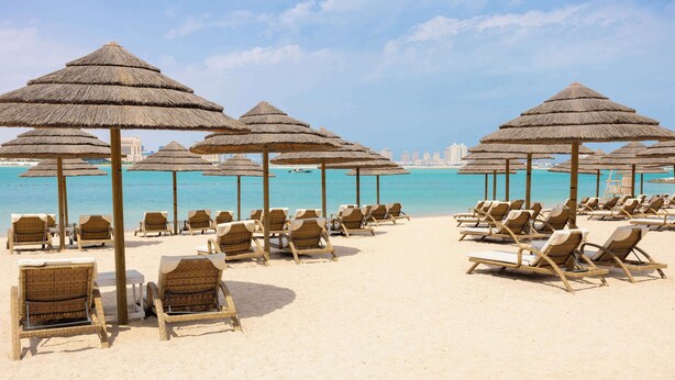 Una playa con hileras de sillones de mimbre y sombrillas de paja, con vista despejada al océano y al horizonte de la ciudad.
