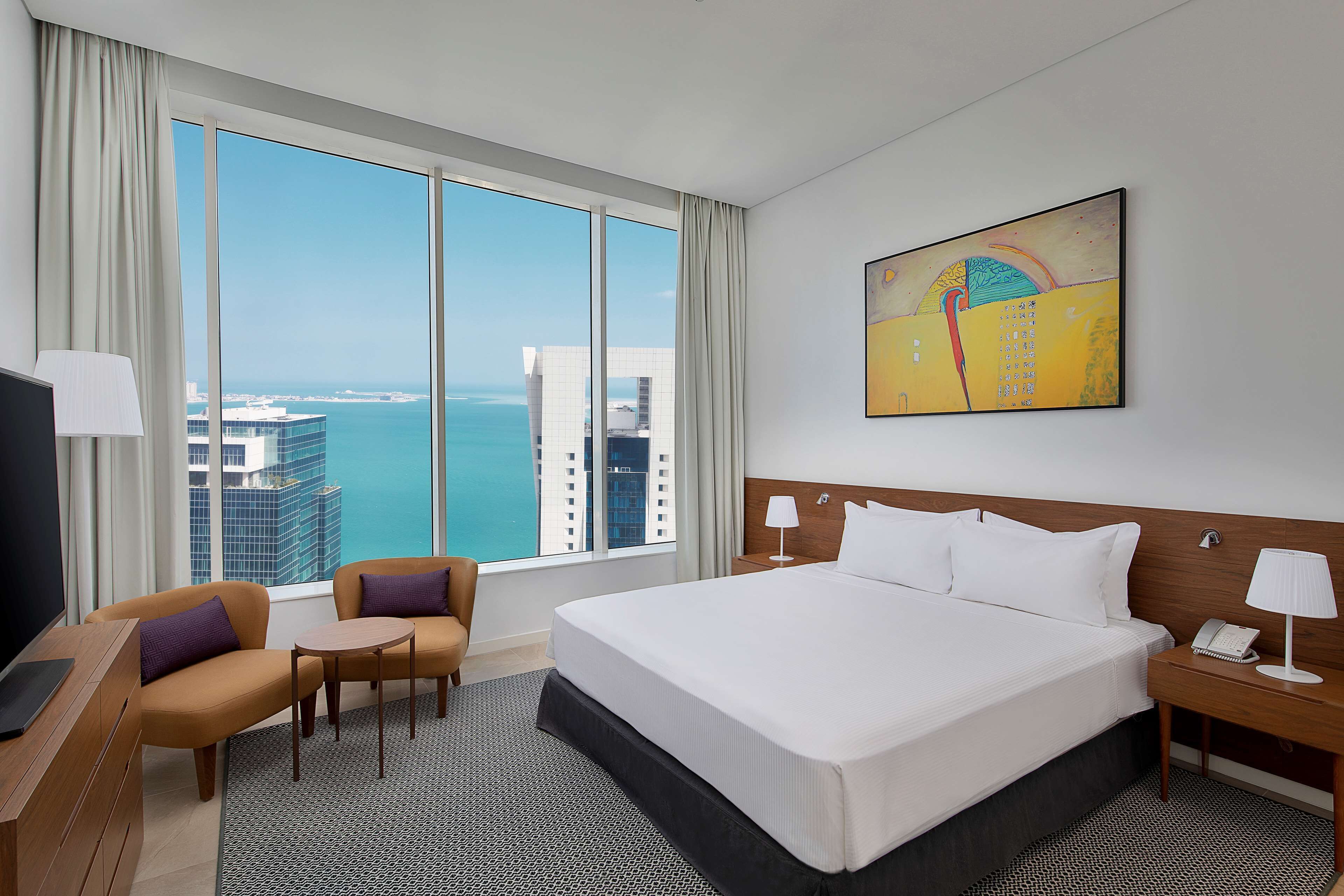 A hotel room with a large bed, a desk with a chair, a TV, and a view of the city and ocean.