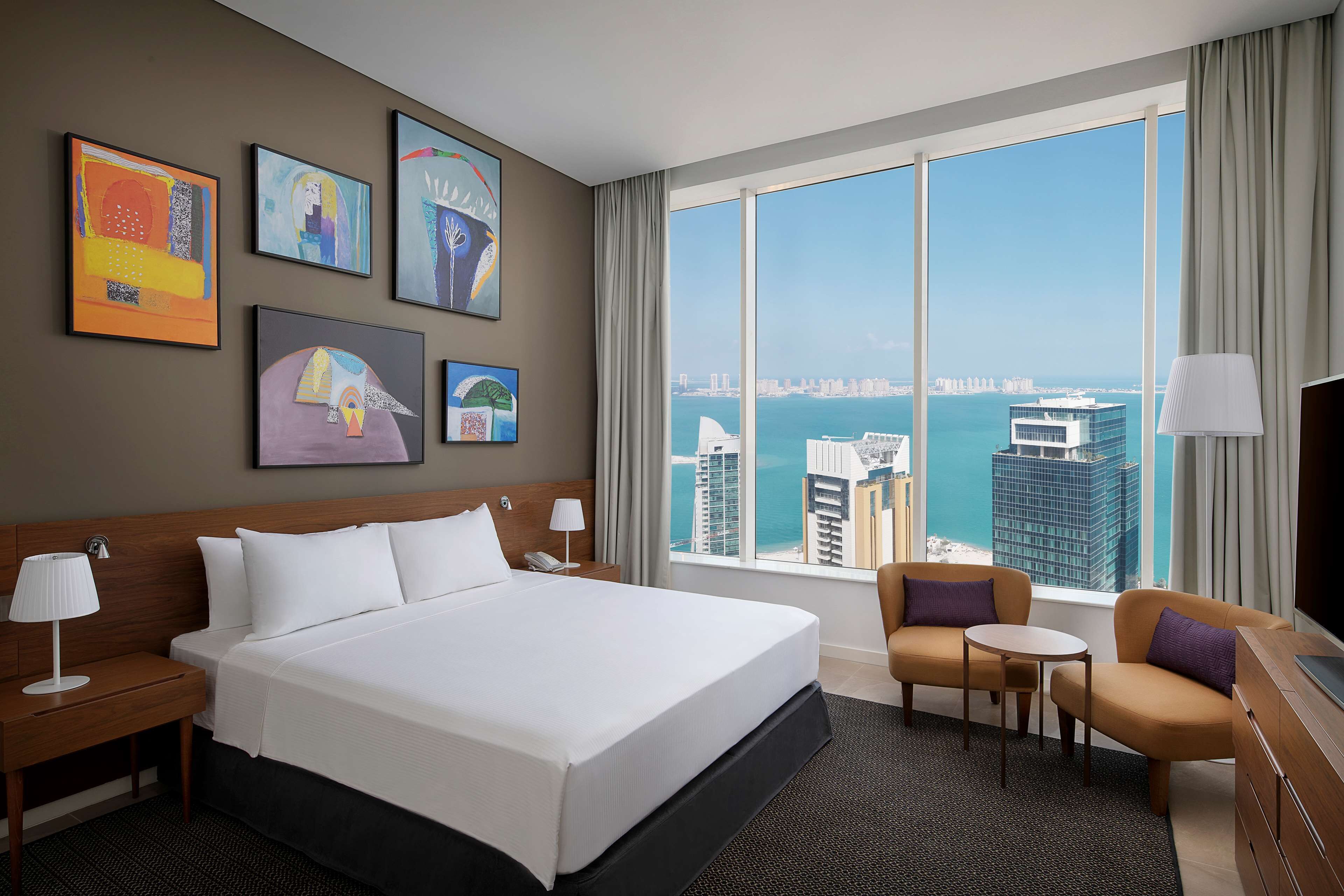 Une chambre d’hôtel avec un grand lit, deux fauteuils, une petite table et un téléviseur. Quatre tableaux ornent le mur et de grandes fenêtres offrent une vue sur le paysage urbain.