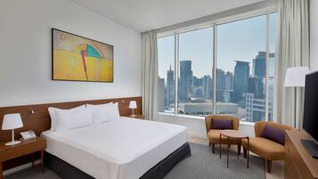 A hotel room with a large bed, a desk with a lamp, a chair, a small table, and a TV. There is a window with a city view.