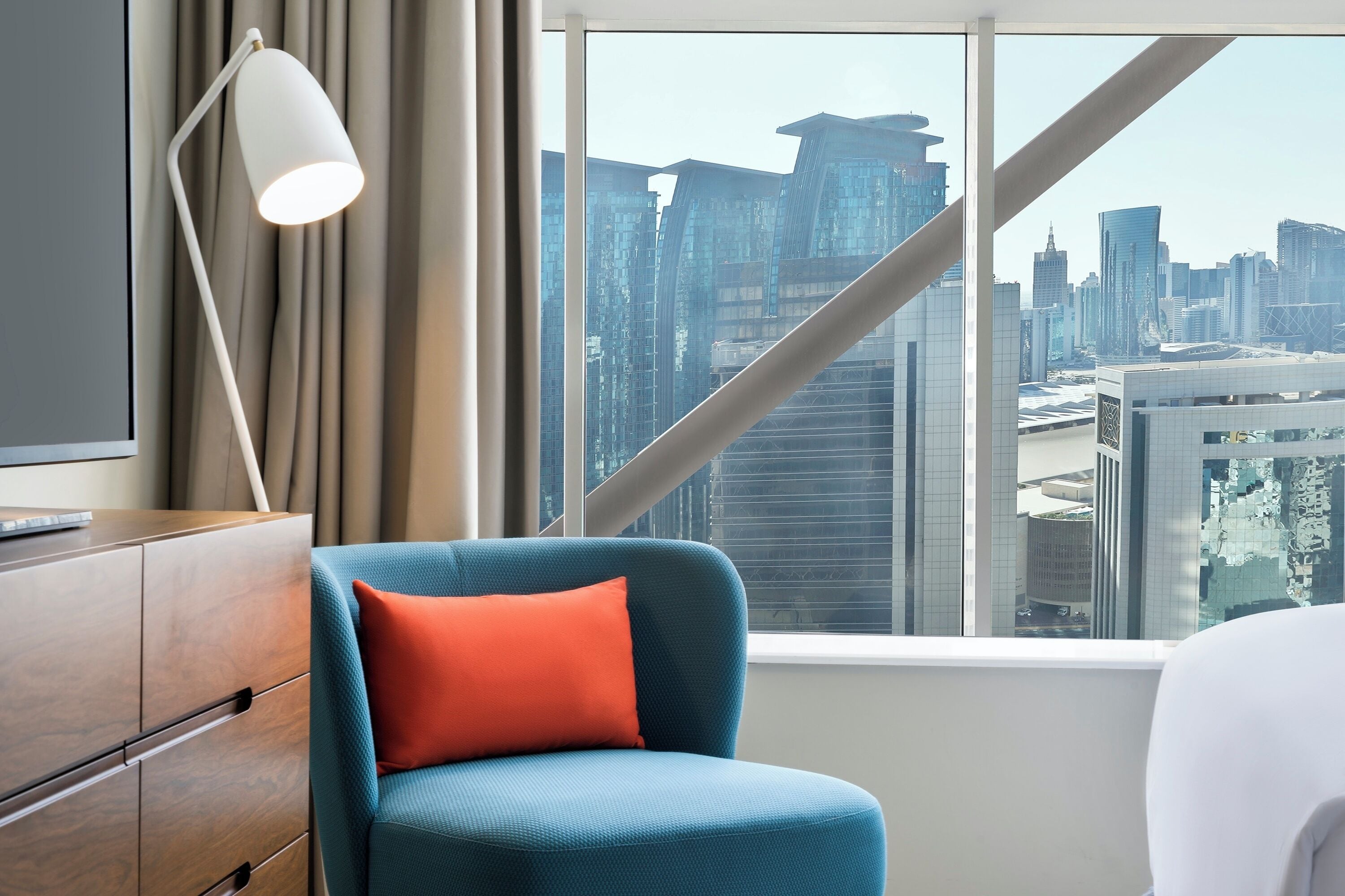 A hotel room with a city view, a blue chair with an orange pillow, a wooden dresser, and a white lamp.