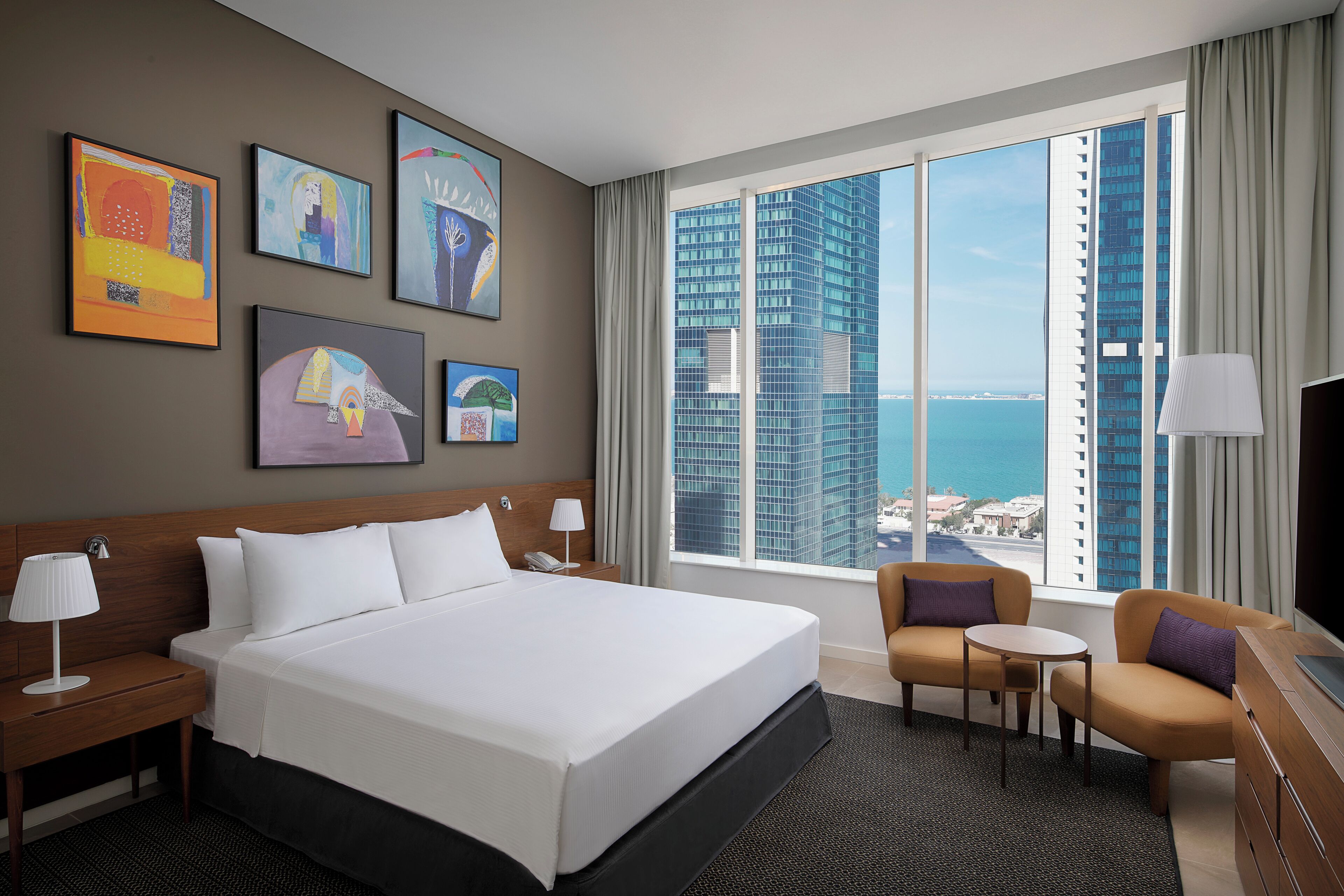 Habitación de hotel con una cama grande, un escritorio, una silla y vistas de un paisaje urbano a través de amplias ventanas.