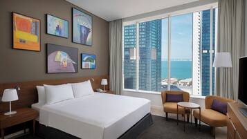 Habitación de hotel con una cama grande, un escritorio, una silla y vistas de un paisaje urbano a través de amplias ventanas.