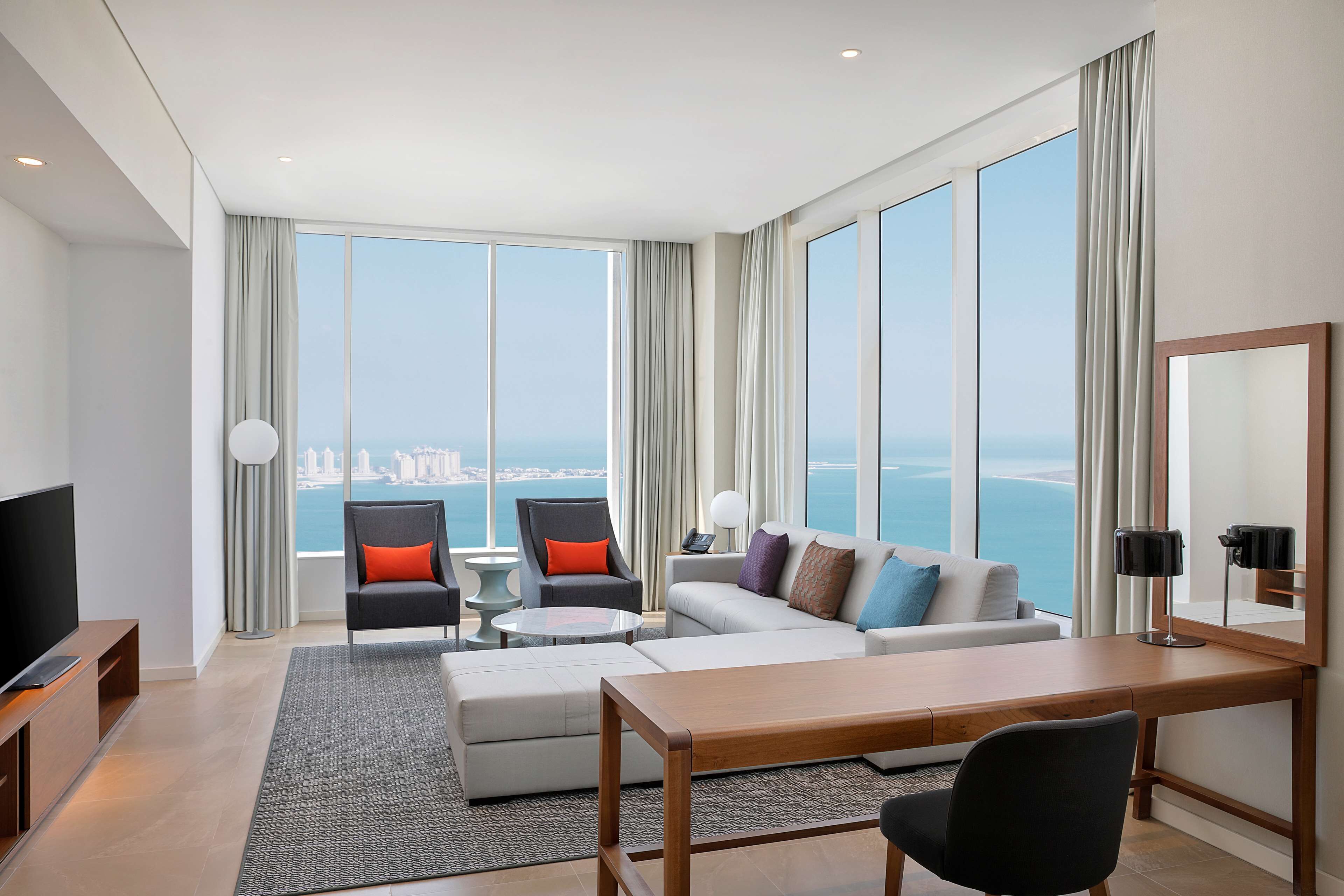A modern hotel room with a large window offering a city view, a flat-screen TV, a sofa, a dining table, and a chair.