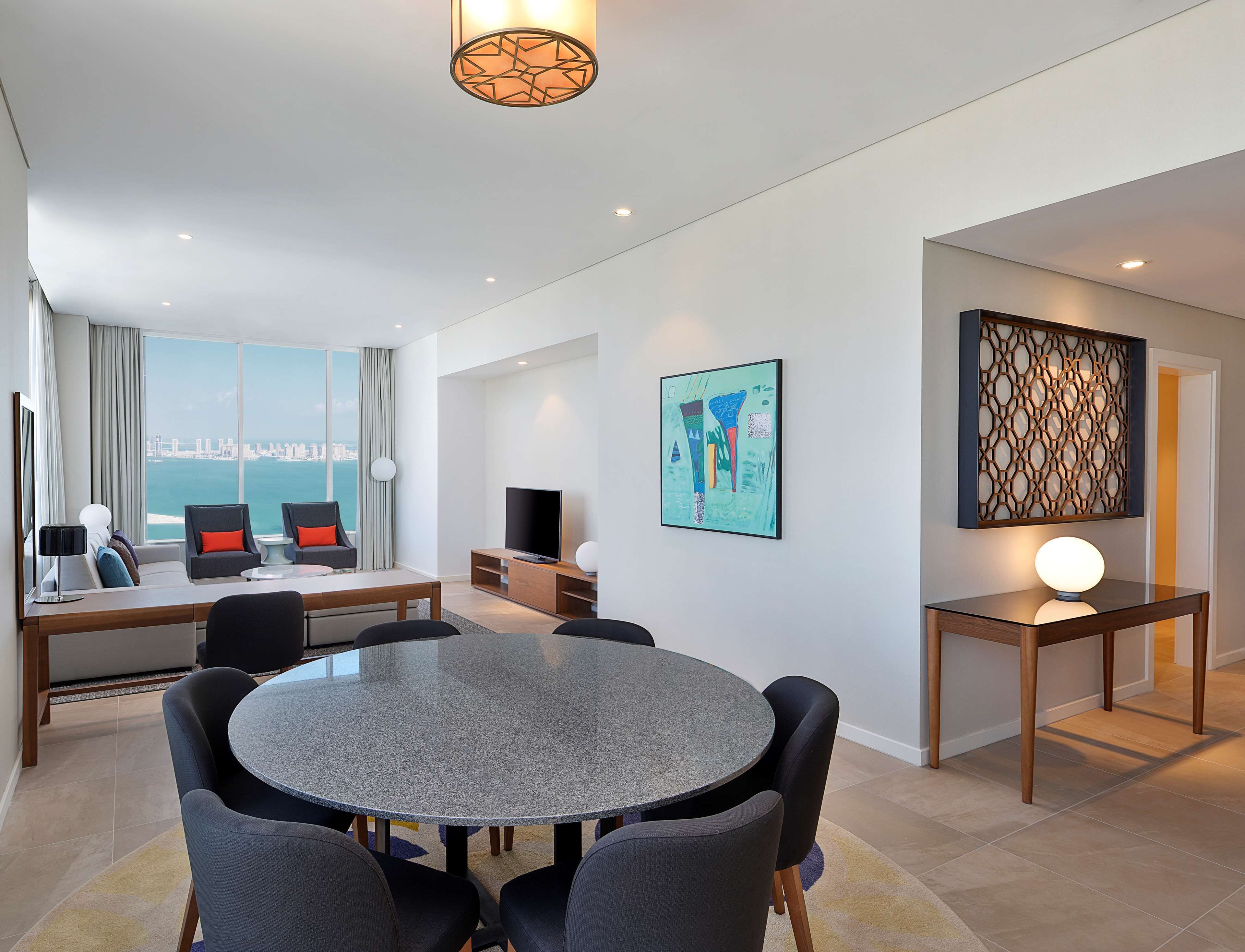 A modern hotel room with a dining area, a TV, and a large window offering a city view.