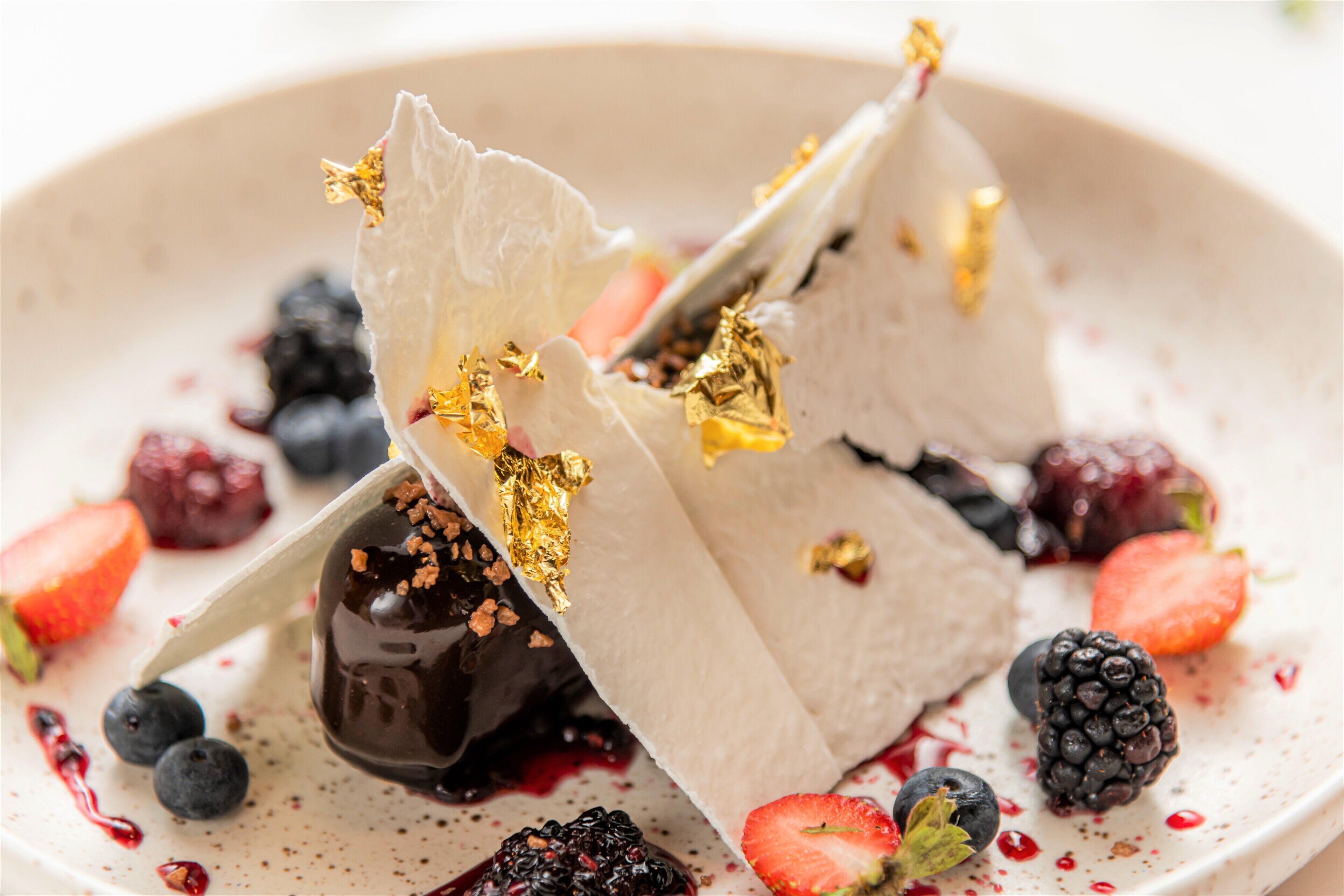 A dessert plate with chocolate-covered strawberries, blueberries, and blackberries, garnished with gold leaf and served on a speckled ceramic plate.
