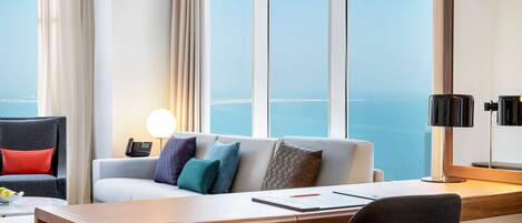 Un hotel moderno con una ventana grande que ofrece vista al mar, un escritorio de madera, un sofá cómodo con cojines de colores y una lámpara en el escritorio.