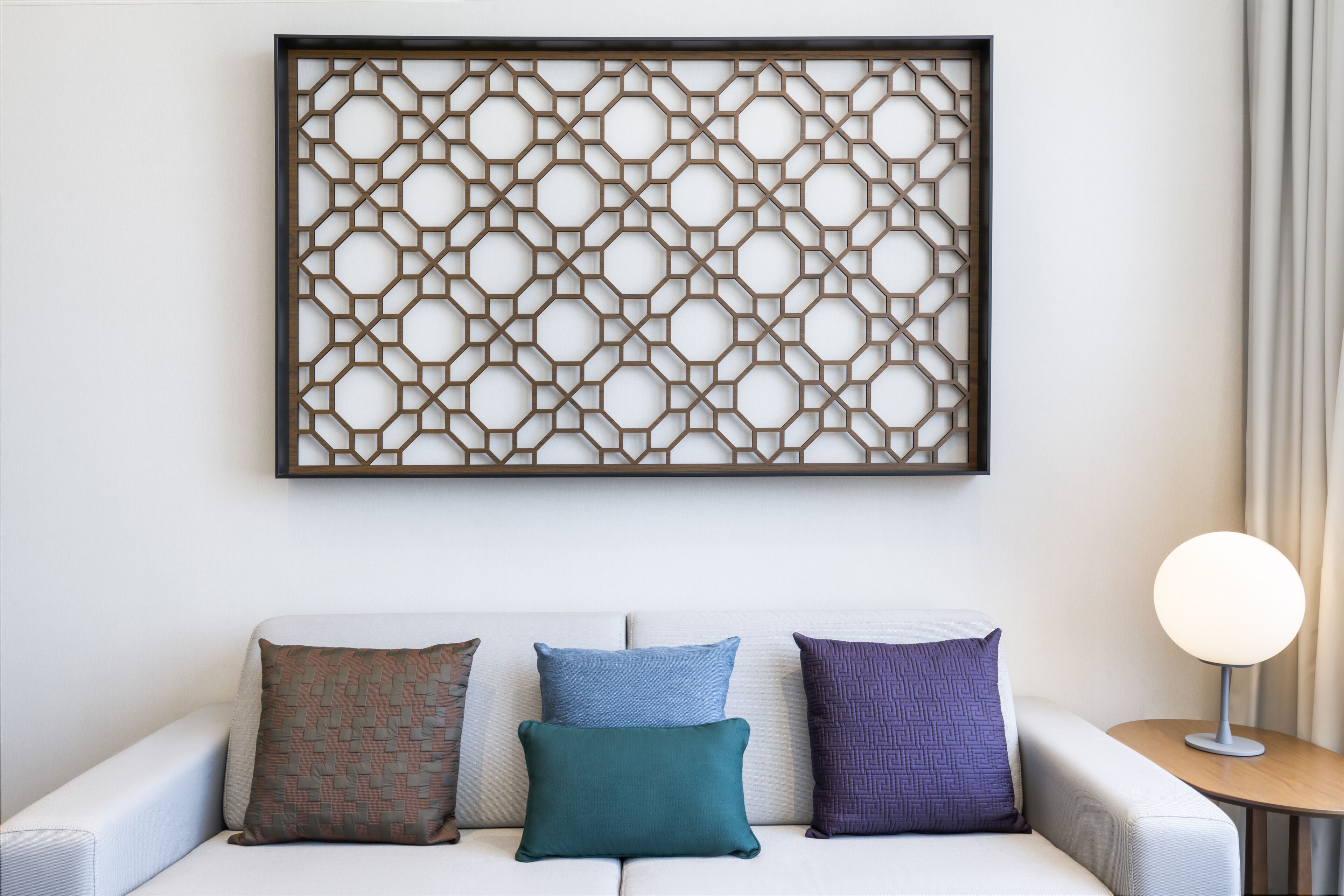 Un soggiorno moderno con un divano bianco, tre cuscini decorati, un quadro a parete geometrico e un tavolino con una lampada.
