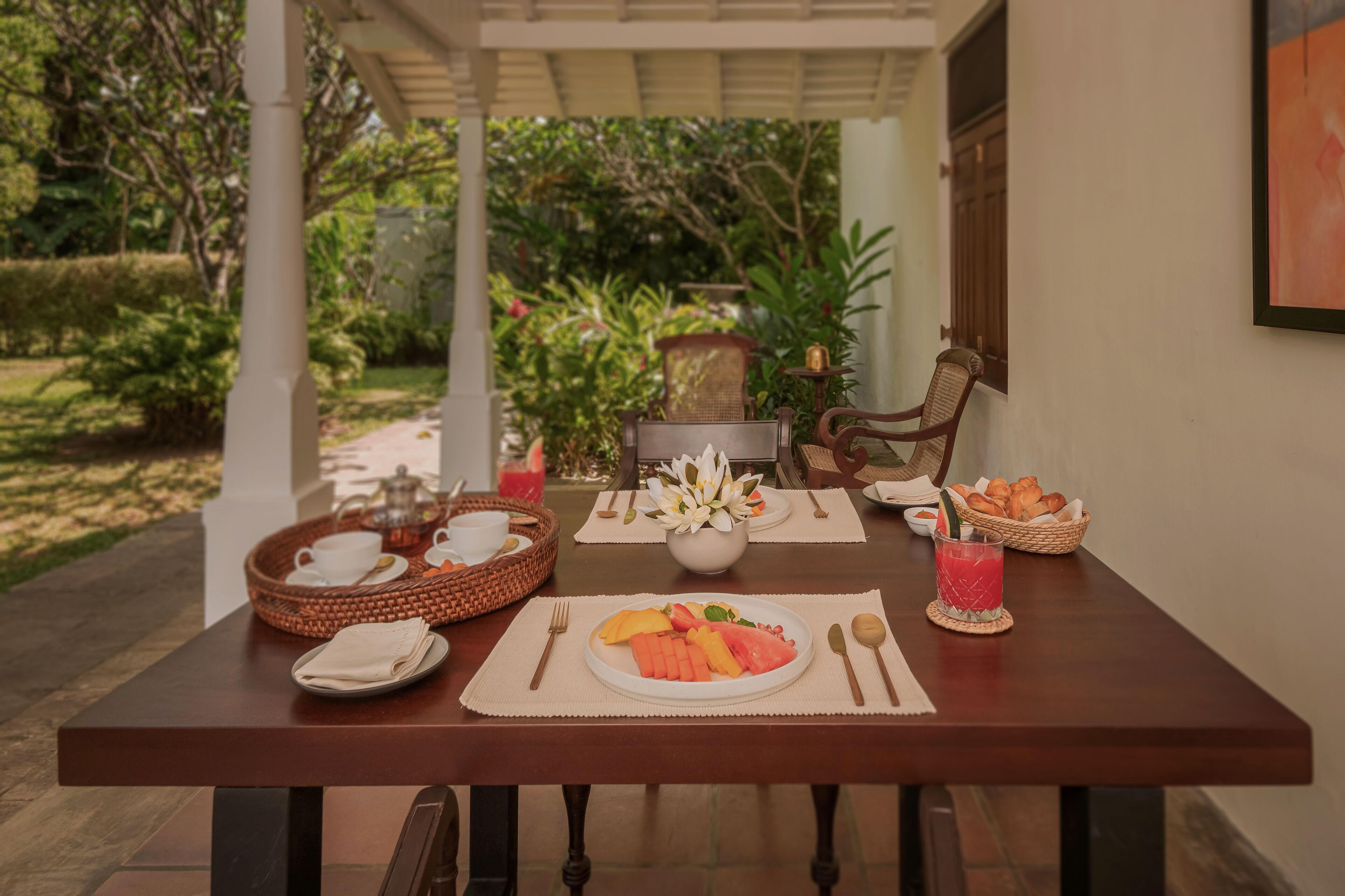 A dining table set with breakfast items, including fruits, bread, and a beverage, in a covered outdoor area with a view of greenery.