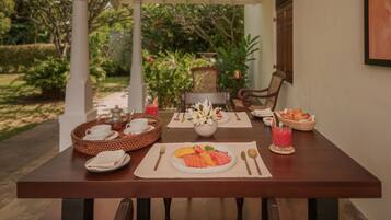 A dining table set with breakfast items, including fruits, bread, and a beverage, in a covered outdoor area with a view of greenery.
