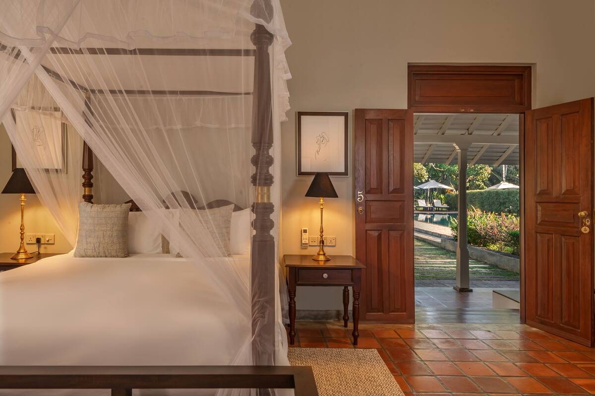 Quarto com cama de dossel, abajures, mesa de cabeceira e vista através de uma porta aberta para área externa com guarda-sóis.