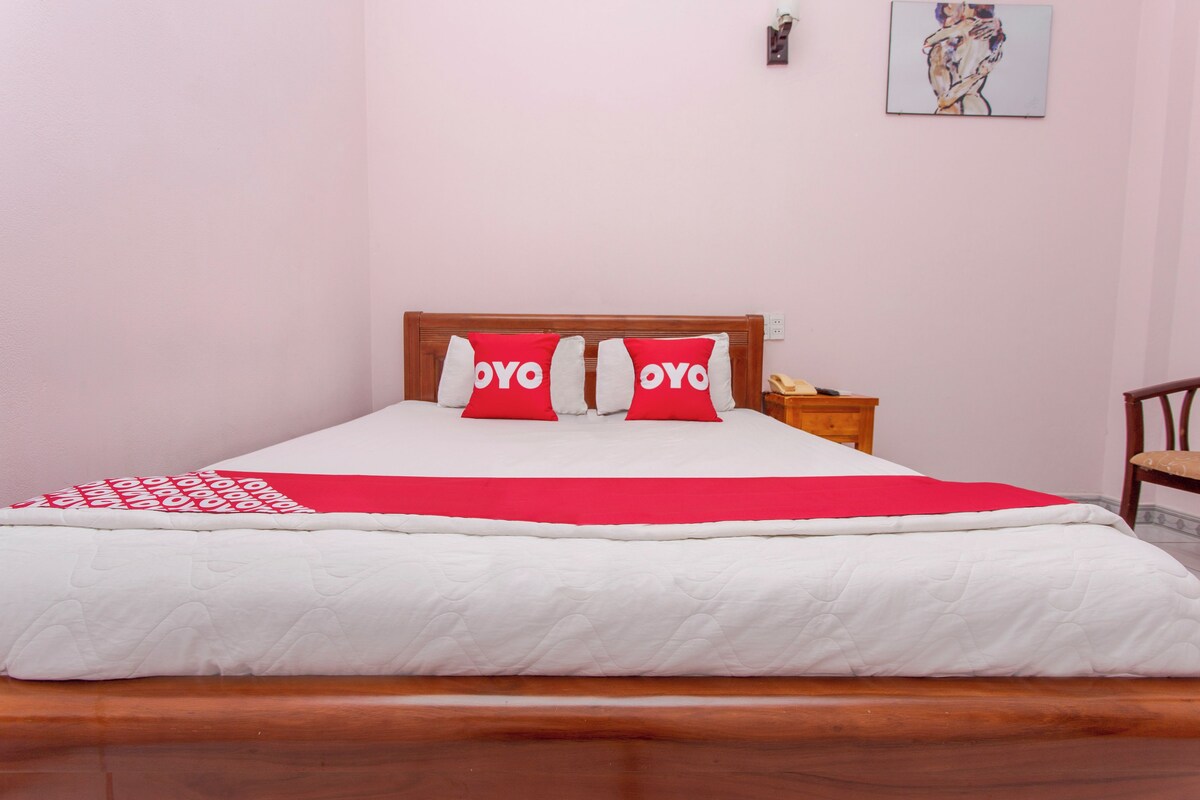 A neatly made bed with a white and red bedspread, two red pillows with 'YOYO' text, a wooden headboard, a nightstand with a lamp, and a painting on the wall.
