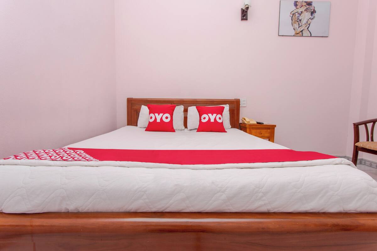 Una cama bien hecha con una colcha roja y blanca, dos almohadas rojas con el texto 'YOYO', un cabecero de madera, una mesita de noche con un teléfono y un cuadro en la pared.