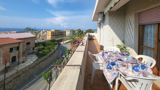 Un balcon avec une table et des chaises, donnant sur une rue et des immeubles, avec un ciel dégagé.
