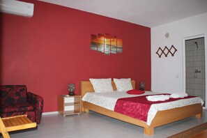 A bedroom with a red wall, a bed with white and red bedding, a wooden nightstand, a red chair, and a wall-mounted artwork.