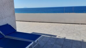 Un balcón con dos sillones azules, piso de baldosas y vista al mar y una montaña lejana.