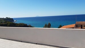 Vista costera desde un balcón con vista al mar, una montaña distante y un edificio pequeño con techo rojo.