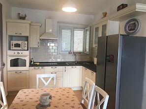 A compact kitchen with white cabinetry, a black refrigerator, a microwave, and a stove. There is a window with blinds, a sink, and a table with chairs. - Apartment Amazing View (Dubrovnik)