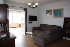 Een woonkamer met een bruine hoekbank, een glazen salontafel, een houten kast, een aan de muur bevestigde televisie en twee ingelijste foto's.